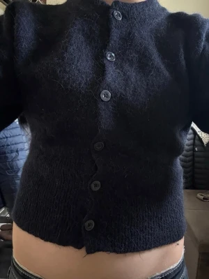 Mörkblå fluffig kofta med knappar - Kort, mörkblå kofta med rund hals och stora knappar framtill. Koftan är stickad i ett fluffigt material och har en croppad passform som ger en trendig look. Perfekt att styla med jeans för en avslappnad outfit.