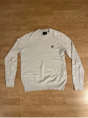 Beige lyle & scott tröja - Beige tröja från Lyle & Scott med rund hals och den klassiska gula örnloggan broderad på bröstet. Tröjan har långa ärmar, ribbade muddar och är tillverkad i ett tunt, mjukt material som passar perfekt till lager på lager. Den är endast använd 1-2 gånger