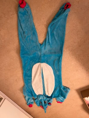 Blå enhörningsdräkt med huva - Rolig blå enhörningsdräkt i mjuk fleece med huva, horn, öron och stora ögon. Dräkten har röda muddar, vit mage och en färgglad regnbågssvans samt man. Perfekt för maskerad eller cosplay och är helkropp med dragkedja framtill.