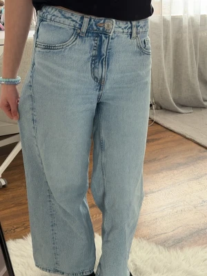 Ljusblåa raka jeans med hög midja - Säljer ett par ljusblå jeans med rak passform och hög midja. Jeansen har klassiska fem fickor, bälteshällor och stängs med knapp och dragkedja. Materialet är kraftig denim i 100% bomull, perfekt för en avslappnad och trendig look.