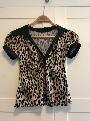 Leopardmönstrad blus med krage - Snygg leopardmönstrad blus med svart krage och korta puffärmar. Blusen har djup V-ringning och svarta detaljer vid ärmslut och att axlar. Perfekt för dig som vill sticka ut med ett djuriskt mönster.