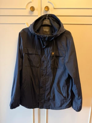 Mörkblå vindjacka från Lyle & Scott - Snygg mörkblå vindjacka från Lyle & Scott med huva och dragsko. Jackan har två bröstfickor med lock, dragkedja och knappstängning samt det klassiska Lyle & Scott-logotypen på bröstet. Tillverkad i lätt och tåligt material, perfekt för blåsiga dagar.