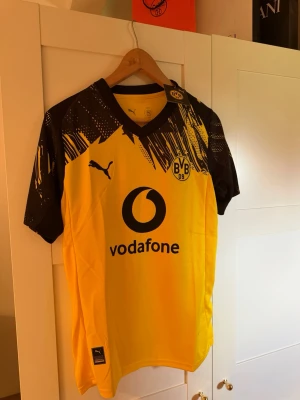 Gul Borussia Dortmund matchtröja Puma - Snygg gul och svart Borussia Dortmund matchtröja från Puma med klubbmärke på bröstet och trycket 'vodafone' på framsidan. Svarta detaljer och grafiskt mönster på axlarna. 'DORTMUND' tryckt på ryggen. Perfekt för fotbollsträning eller att bära till vardags.