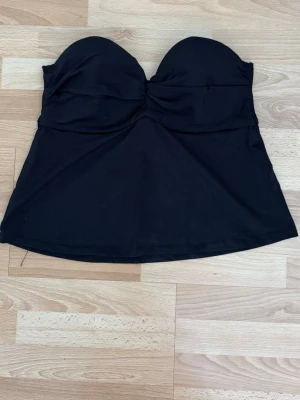 Svart bandeau tankini topp utan knytning - Snygg svart bandeau bikini topp med vadderad byst och lätt rynkning framtill. Modellen har en kjolliknande nederdel som ger en retro känsla. Tillverkad i stretchigt material som passar perfekt för strand eller pool. Storleklapp bortklippt, men den är xs/s, under brösten 72cm.