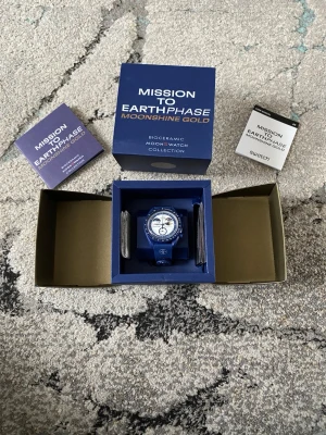 Omega x Swatch MoonSwatch Moonshine Gold - Säljer en blå Omega x Swatch Mission to Earthphase Moonshine Gold MoonSwatch. Klockan har en rund boett i biokeramik, vit urtavla med blå och guldiga detaljer samt blå textilrem. Kommer i originalförpackning med tillhörande manualer.