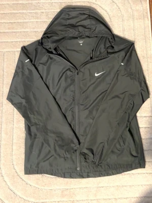 Grå Nike miler windbreaker - Snygg grå regnjacka från Nike med dragkedja framtill och justerbar huva. Jackan har diskreta reflexdetaljer på ärmarna och Nike-logga på bröstet. Tillverkad i lätt, vattenavvisande material som passar perfekt för regniga dagar.