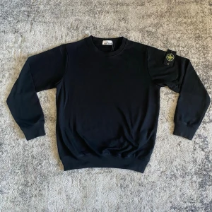 Svart stickad tröja från Stone Island - Snygg svart stickad tröja från Stone Island med klassisk rund halsringning och ribbade muddar vid ärmslut och nederkant. Tröjan har den ikoniska Stone Island-loggan på vänster ärm. Perfekt för dig som gillar stilrena plagg.