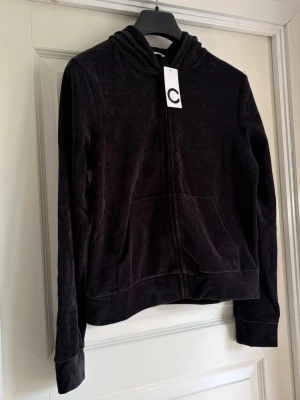 Svart velourhoodie från Cubus - Mjuk och skön svart hoodie i velour från Cubus. Hoodien har dragkedja framtill, två fickor och en klassisk huva. Perfekt för en chill och avslappnad stil. Materialet är lent och ger en lyxig känsla.