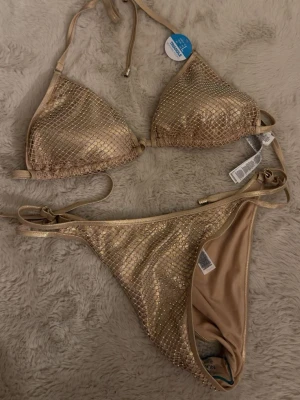 Glittrig guldig bikini från newyorker - Supersnygg trekantsbikini i guld från Hunkemöller med glittrigt mönster och knyt i både topp och trosa. Perfekt för sommarens strandhäng och poolpartyn. Materialet är mjukt och följsamt syntet som sitter skönt mot huden. Storlek L men passar mer som M