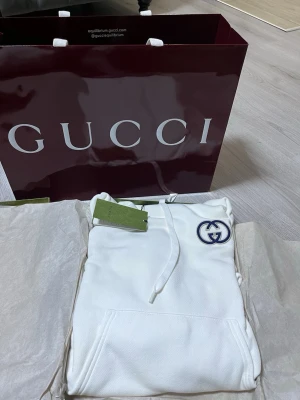 Vit hoodie från Gucci med logga - Snygg vit hoodie från Gucci med klassisk blå logga broderad på bröstet. Hoodien har huva med dragsko och en stor magficka framtill. Tillverkad i mjukt bomullsmaterial som känns lyxigt och bekvämt. Perfekt för dig som vill ha en stilren och exklusiv look.