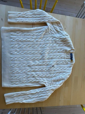 Krämvit kabelstickad tröja Polo Ralph Lauren - Kabelstickad vit tröja från Polo Ralph Lauren med rund halsringning och diskret broderad logga på bröstet. Tröjan har långa ärmar och ribbade muddar vid ärmslut och nederkant. Perfekt för en klassisk och stilren look.