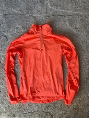 Neonorange träningströja Swedemount - Säljer en neonorange långärmad träningströja från Swedemount med halv dragkedja framtill och hög krage. Perfekt för löpning eller utomhusträning. Tillverkad i lätt och snabbtorkande material som andas. Snygg och funktionell för aktiva dagar.