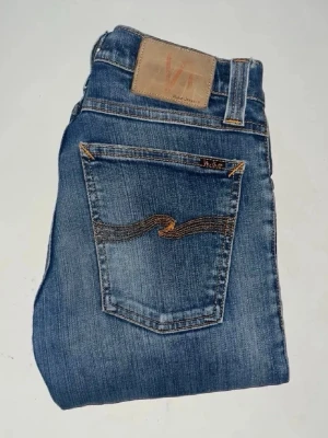 Nudie jeans  - Säljer ett par klassiska blå skinny jeans från Nudie Jeans, i modellen tajht long john. Jeansen har snygga slitningar och kontrastsömmar. Midja 33cm, längd 106cm. JAG SKICKAR EJ FLER BILDER NÄR JAG BÄR JEANSEN! för bättre inblick i passformen rekommenderar jag att googla på modellnamnet. För storleksguide kolla måtten noggrant då jeansen alltid kan vara uppsydda heller krympta och jag ej tar emot returer :) s5,2