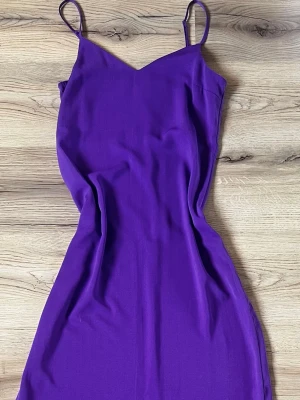 Lila långklänning med smala axelband - En snygg lila långklänning med smala axelband och v-ringad hals. Klänningen är ärmlös och har en figurnära passform som framhäver siluetten. Tillverkad i ett mjukt och slätt material som ger ett elegant intryck.