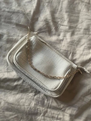 Vit croco handväska med kedja - Snygg vit handväska i croco-mönstrat konstläder med en kort kedja i metall. Väskan har två fack och dragkedja upptill. Perfekt accessoar för att lyfta din outfit med en trendig touch.