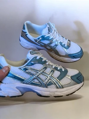 Asics Gel-1130 vita och blå sneakers - Snygga Asics Gel-1130 sneakers i vitt med blå och silver detaljer. Skorna har mesh-överdel för bra ventilation och klassisk Asics-logga på sidan. Lågt skaft och robust sula som ger grym komfort. Perfekta för dig som gillar sportig stil.