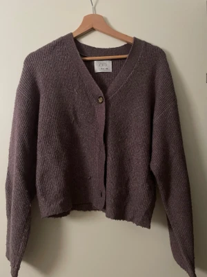 Lila ribbad kofta från Zara - Mysig lila kofta från Zara med ribbad struktur och v-ringning. Koftan har en avslappnad passform, långa ärmar och stängs med en knapp framtill. Perfekt att slänga över en t-shirt för en chill look. Passar för de som har Xs, S och M