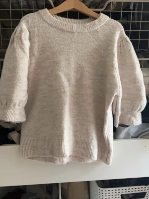 Beige stickad tröja med puffärm - Mysig beige stickad tröja med rund halsringning och trendiga puffärmar. Tröjan har ribbade muddar vid ärmslut och nederkant, vilket ger en snygg och avslappnad look. Perfekt för dig som gillar stilrena och bekväma plagg.