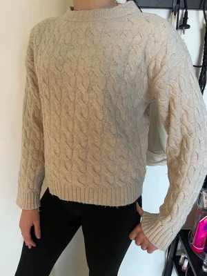 Beige kabelstickad tröja - Kabelstickad tröja i beige med rund hals och långa ärmar. Tröjan har ribbade muddar vid ärmslut och nederkant för en clean look. Materialet ser mjukt och mysigt ut, perfekt för lager på lager under kalla dagar.