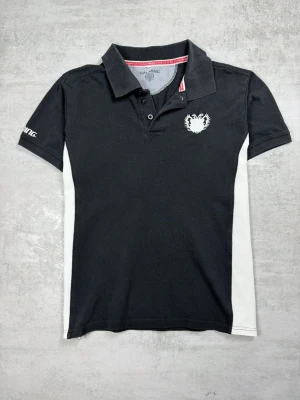 Svart pikétröja från Salming - Klassisk svart pikétröja från Salming med vita sidopaneler och broderad logga på bröstet. Tröjan har krage, knappar framtill och korta ärmar. Perfekt för en sportig och avslappnad stil.