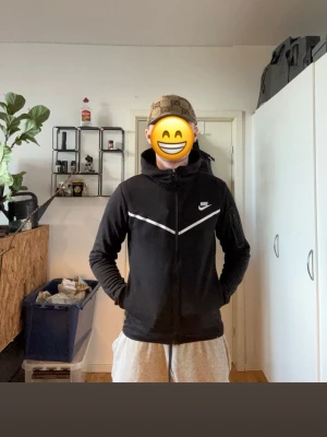 Svart Nike hoodie med dragkedja - Säljer en svart hoodie från Nike med vit logga på bröstet och vita detaljer över bröstet. Hoodien har dragkedja framtill, huva och fickor på sidorna. Perfekt för dig som gillar sportig och clean stil. Materialet känns mjukt och bekvämt.
