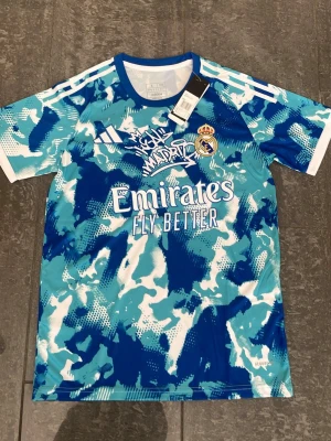 Real Madrid Jersey - Snygg Real Madrid träningströja från Adidas i blått, turkost och vitt camomönster. Tröjan har klubbmärke och Adidas-logga på bröstet samt texten 'Emirates Fly Better' framtill. Tillverkad i lätt och ventilerande syntetmaterial, perfekt för träning.