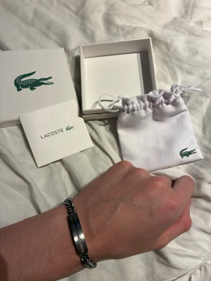 Silverfärgat armband från Lacoste - Snyggt silverfärgat armband från Lacoste med en stilren platta där Lacoste-loggan är ingraverad. Armbandet har en kedjedesign i metall och kommer med originalförpackning och tygpåse med Lacoste-krokodilen på. Perfekt accessoar för en clean och modern look. Nypris cirka 1200kr..