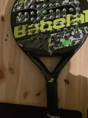 Babolat padelracket Rival svart/gul - Padelracket från Babolat, modell Rival, i svart med gula detaljer och kamouflagemönster. Racketen har rund form, många hål och greppvänligt handtag. Tillverkad med carbon frame för extra hållbarhet och prestanda.
