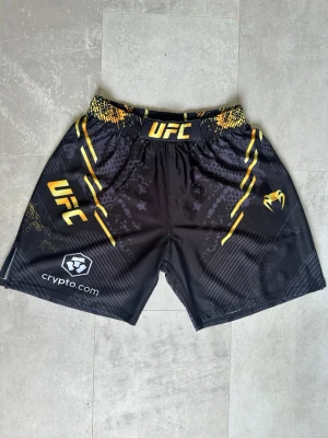 shorts till kampsport (Venom) - Snygga svarta fightshorts från Venum med UFC-logga och gula detaljer. Du kan använda de till MMA, Muay Thai, Boxning och fighting. De passar perfekt för träning och kampsport. De är helt nya utan lapparna. Passar även dig som har storlek S