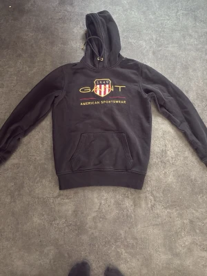 Svart hoodie från GANT med tryck - Säljer en svart hoodie från GANT med huva och dragsko. På bröstet finns ett stort tryck med GANT-logga, amerikansk sköld och texten 'American Sportswear'. Hoodien har magficka och ribbade muddar. Perfekt för en avslappnad och sportig stil. 