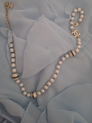 pärlhalsband - Säljer ett elegant halsband  med vita pärlor och guldfärgade detaljer. Halsbandet har flera klassiskaloggor i metall och några större pärlor med svarta och guldfärgade ränder. Perfekt accessoar för att lyfta din outfit.