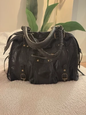 Handväska vintage - Cool svart handväska i skinnimitation med detaljer som nitar, spännen och flera dragkedjor. Väskan har dubbla handtag och en mjuk, lite slouchy form. Ena dragkedjan sönder annars bra skick!