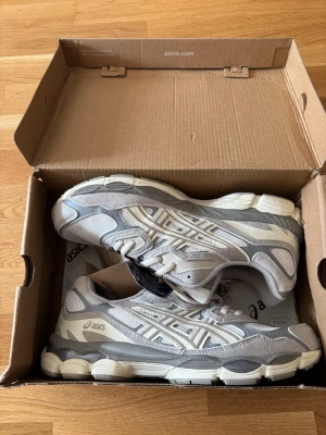 Gråvita sneakers från Asics - Snygga sneakers från Asics i grått och vitt med mesh och mockadetaljer. Skorna har en chunky sula och klassisk Asics-logga på sidan. Perfekta för dig som gillar sportig och trendig stil. Lågt skaft och snörning framtill.