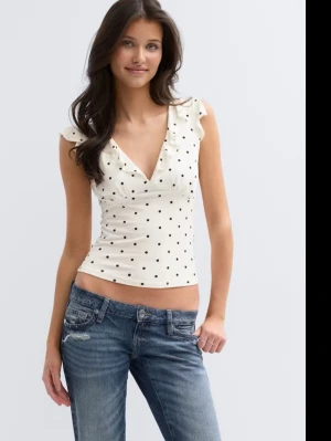 Vit prickig croppad topp - Snygg vit croppad topp med svarta prickar och v-ringning. Toppen har volangdetaljer vid axlarna och är ärmlös, vilket ger en fräsch och trendig look. Perfekt för dig som vill sticka ut lite extra i sommar.