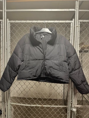 Svart cropped pufferjacka H&M Divided - Säljer en svart cropped pufferjacka från H&M Divided. Jackan har hög krage, dragkedja framtill och två stora fickor. Perfekt för kalla dagar och ger en cool, avslappnad vibe. Materialet är syntetiskt och jackan är vadderad för extra värme.