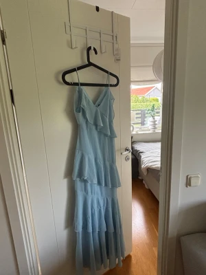 Ljusblå långklänning med volanger - Säljer en ljusblå långklänning med tunna axelband och v-ringning. Klänningen har flera lager volanger och en luftig, flowy passform som passar perfekt till sommaren. Materialet känns lätt och svalt, perfekt för varma dagar.