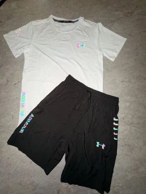 Vit t-shirt från Underarmour - Snygg vit t-shirt med regnbågsfärgade loggor och textdetaljer från Under Armour. T-shirten har rund hals, korta ärmar och är tillverkad i ett mjukt material som passar perfekt till träning eller chill. Stilsäker och modern look med färgglada accenter.