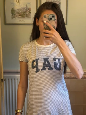 Randig GAP t-shirt med logga - Säljer en vit och grårandig t-shirt från GAP med stor mörkblå logga framtill. T-shirten har korta ärmar, rund halsringning och är gjord i mjuk bomull. Perfekt för en avslappnad och stilren look.