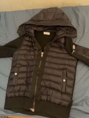 Svart dunväst med huva från Moncler - Snygg svart dunväst från Moncler med huva och dragkedja framtill. Västen har två fickor med tryckknappar och Moncler-logga på ärmen. Perfekt för lager-på-lager och streetstyle.
