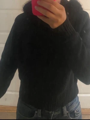 Svart stickad hoodie med luva - Säljer en jättefin svart stickad kofta med dragkedja. Den har en mysig, stor huva med mjuk fuskpäls och ribbade detaljer vid ärmsluten och nedtill. Tröjan är i ett skönt och varmt material. Mycket fint skick! 