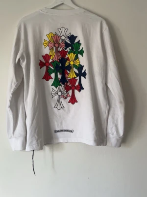 Vit långärmad topp från Chrome Hearts - Vit långärmad topp från Chrome Hearts med färgglada kors på ryggen och mindre tryck på bröstfickan. Toppen har en avslappnad passform och är tillverkad i mjuk bomull. Perfekt för dig som gillar streetwear och unika detaljer.