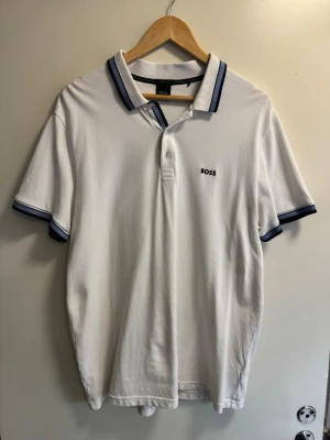 Vit pikétröja från BOSS - Klassisk vit pikétröja från BOSS med blå detaljer på krage och ärmslut. Tröjan har korta ärmar, knappslå framtill och diskret BOSS-logga broderad på bröstet. Tillverkad i mjuk bomull för en bekväm känsla. Står XXL Men passar även XL