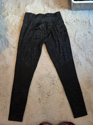 Svarta mönstrade leggings Newbody - Säljer ett par svarta leggings från Newbody med diskret mönster och hög midja. Materialet är stretchigt och bekvämt, perfekt för träning eller chill. Paneler i benen ger en snygg kontrast och extra stilpoäng. Tight passform och elastisk midja.