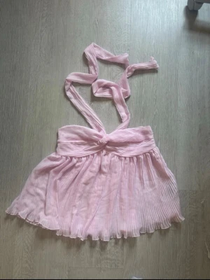 Ljusrosa halter topp från Gina Tricot - Supersöt ljusrosa halterneck topp från Gina Tricot med plisserad, luftig design och knytband i halsen. Perfekt för dig som vill ha en feminin och trendig look. Toppen är ärmlös och har en flowy känsla. Endast använd en gång