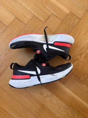 Nike React Miler – Löparskor – Strl 41 – Svart/Vit/Röd - Säljer ett par svarta Nike löparskor med vita och röda detaljer. Skorna har en tjock, vit sula med struktur och svart yttersula. Ovandelen är i mesh för bra andningsförmåga och har tydliga vita swoosh-loggor på sidorna. Snygga och sportiga sneakers som sticker ut.