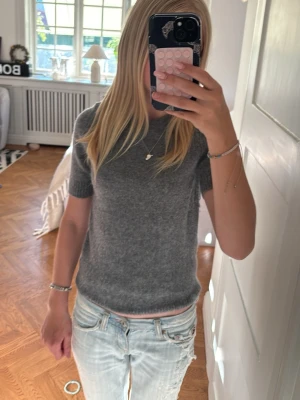 Grå stickad t-shirt från Zara - Mysig grå stickad t-shirt från Zara med korta ärmar och rund halsringning. Tröjan har en enkel och stilren design i mjukt material, perfekt att matcha med jeans eller kjol för en avslappnad look.