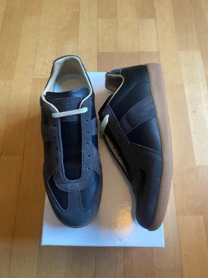Margiela Gats - (Helt Nya) Snygga sneakers med detaljer i mocka och slät läderlook. Skorna har beige innersula och yttersula i gummi. Klassisk låg modell med vita skosnören och stilren design som passar till det mesta.