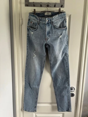 Rebel Ljusblå raka jeans med slitningar - Snygga ljusblå jeans med raka ben och slitna detaljer på låret och vid fickorna. Klassisk femficksmodell med hög midja och dragkedja. Perfekta för en avslappnad och trendig look.