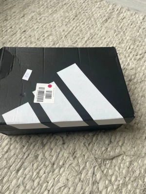 Adidas Predator vita fotbollsskor - Snygga vita Adidas Predator fotbollsskor med silvergrå detaljer och blå accenter. Skorna har klassisk snörning och är tillverkade i syntetmaterial med mönstrad ovandel för extra grepp. Perfekta för dig som vill sticka ut på planen.