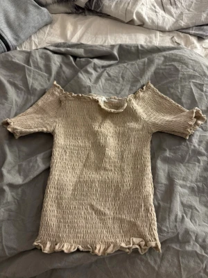 Beige offshoulder topp med volang - Trendig beige offshoulder topp med korta ärmar och volangdetaljer vid halsringning och nederkant. Smockad och stretchig för en tight passform, perfekt till sommaren. Enkel att matcha med både jeans och kjol.